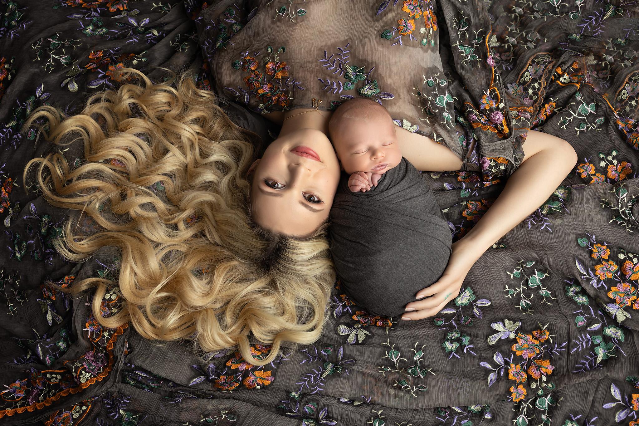 Stunning mother laying down with her newborn baby wrapped in gray and her blonde curls splayed out