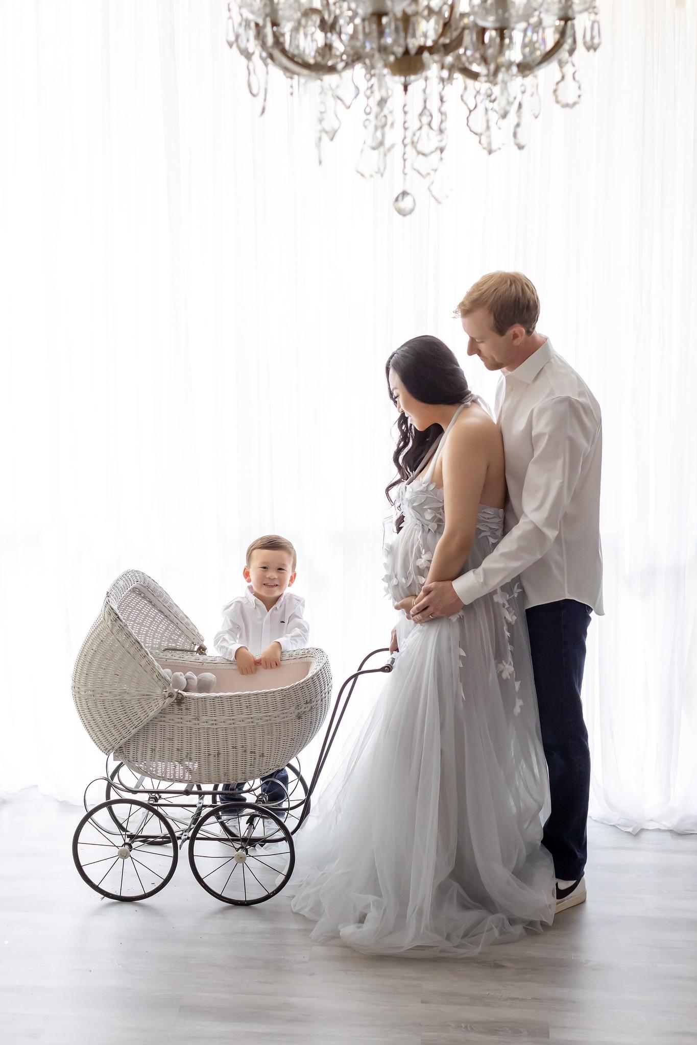 Family of three including pregnant mom wearing a gorgeous tulle gown embraced by dad and looking down at a vintage stroller with their son standing behind the stroller and smiling excitedly at the camera.