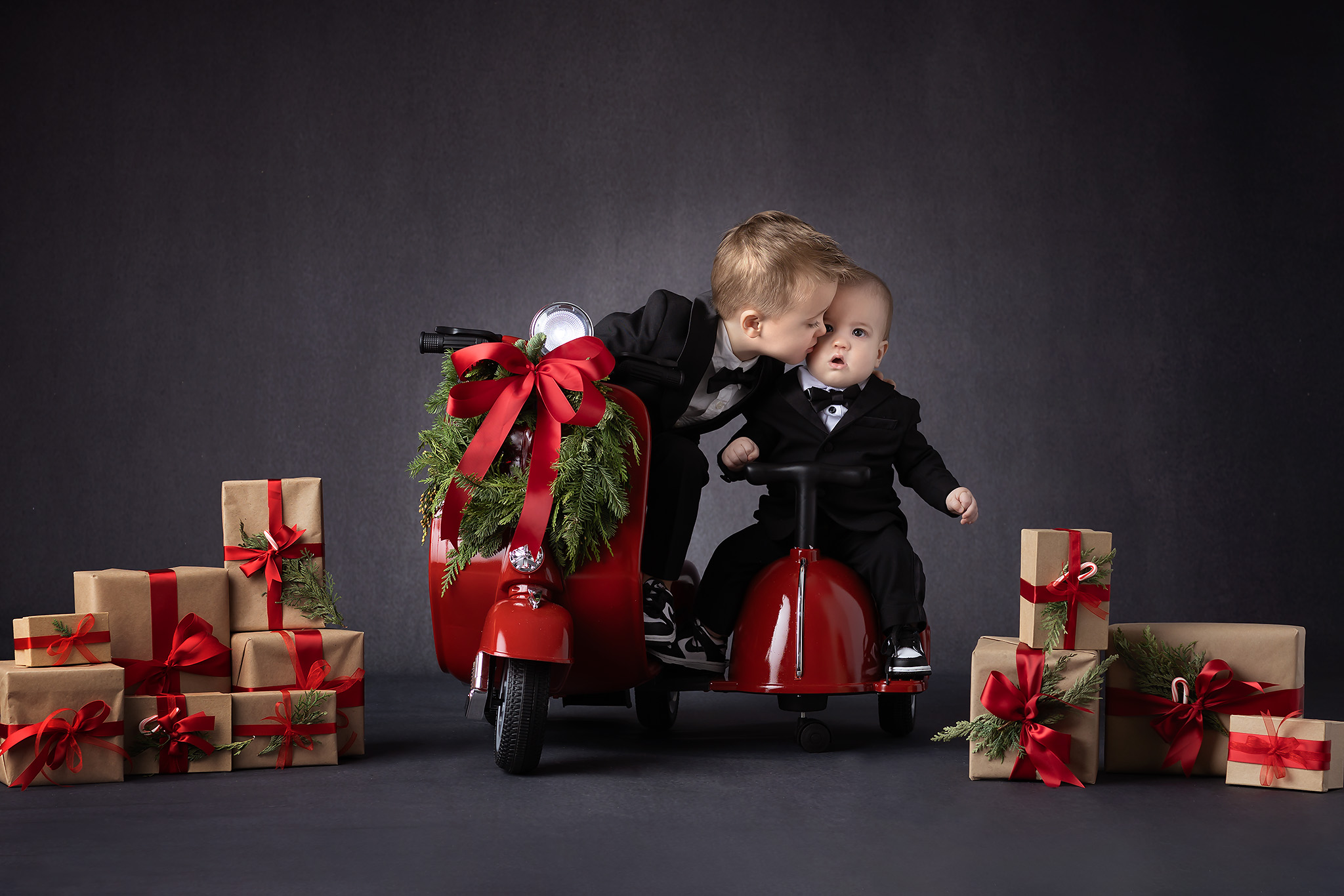 Photo of older brother kissing little brother's cheek while sitting on little scooters surrounded by Christmas holiday packages.