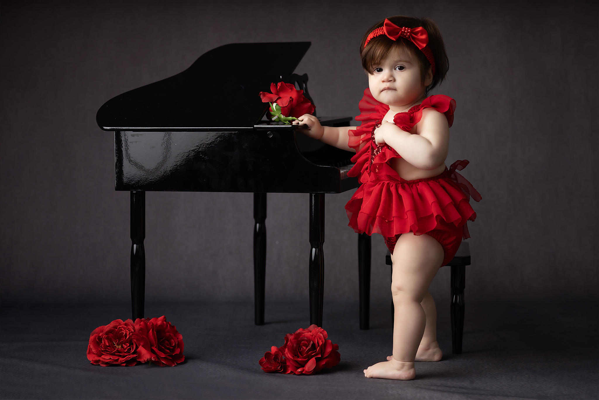 An infant girl in a red dress leans on a black piano in a studio after meeting babysitters in Dallas
