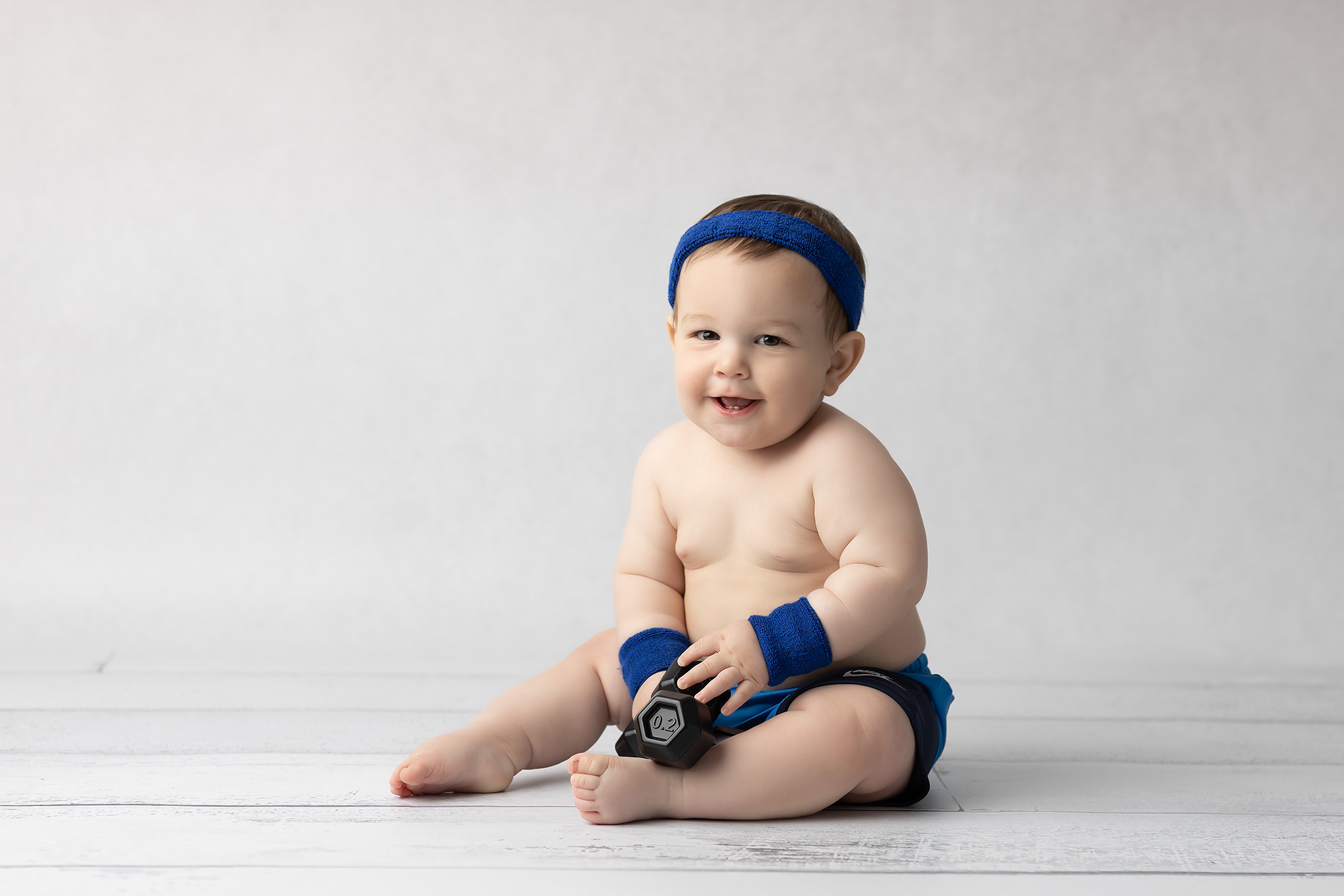 A smiling infant sits on wood floor in sweat bands holding a tiny weight after meeting babysitters in Dallas