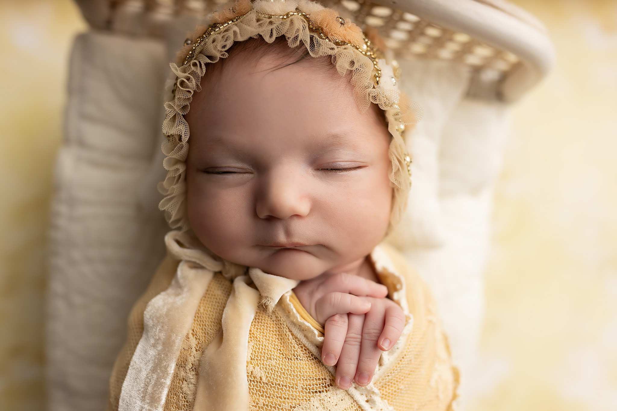 A closeup of a newborn baby sleeping in a bonnet and swaddle on a tiny bed in a studio