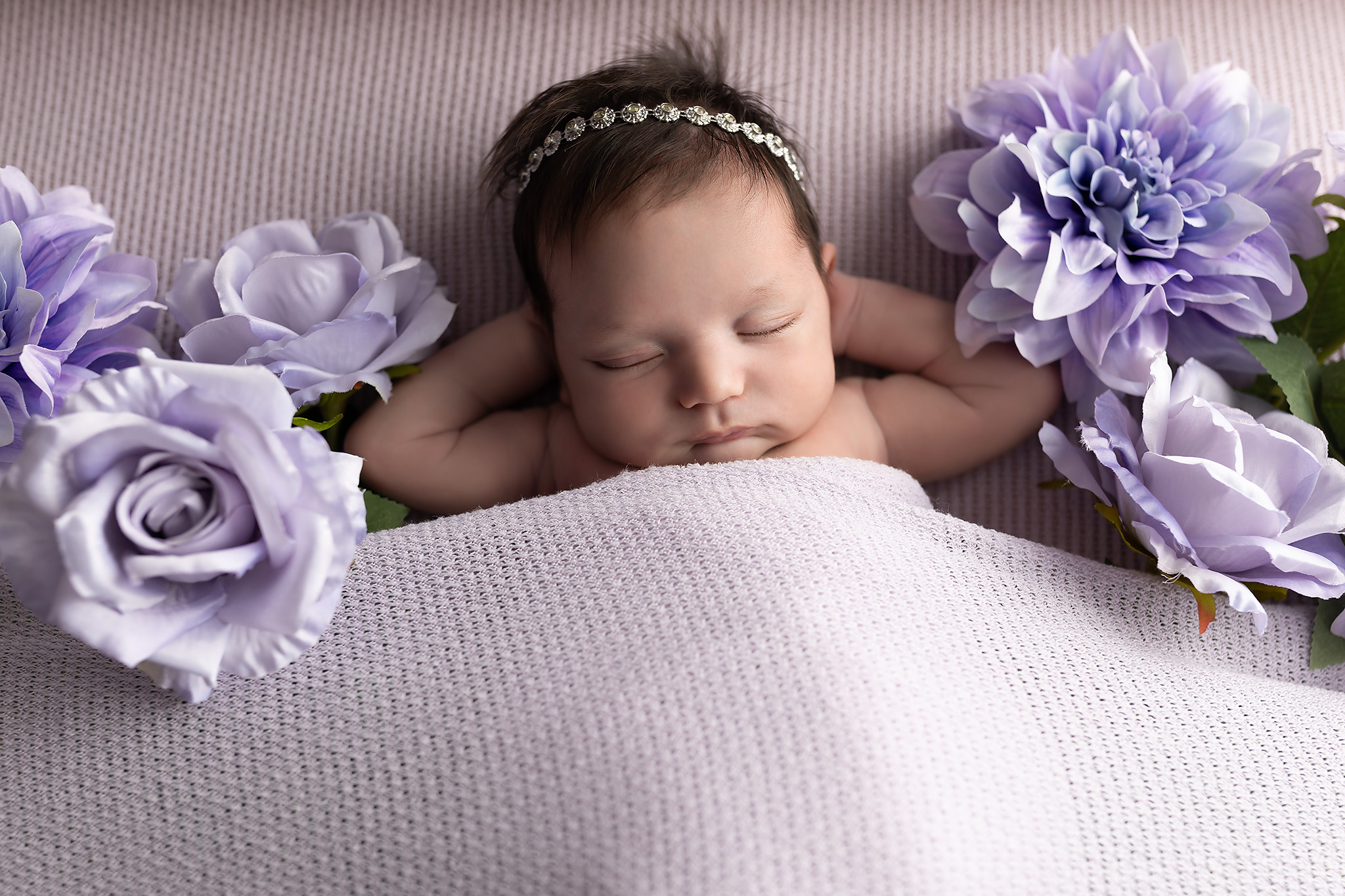 A sleeping newborn girl with hands behind her head surrounded by flowers tucked under a blanket after meeting sleep consultants in fort worth