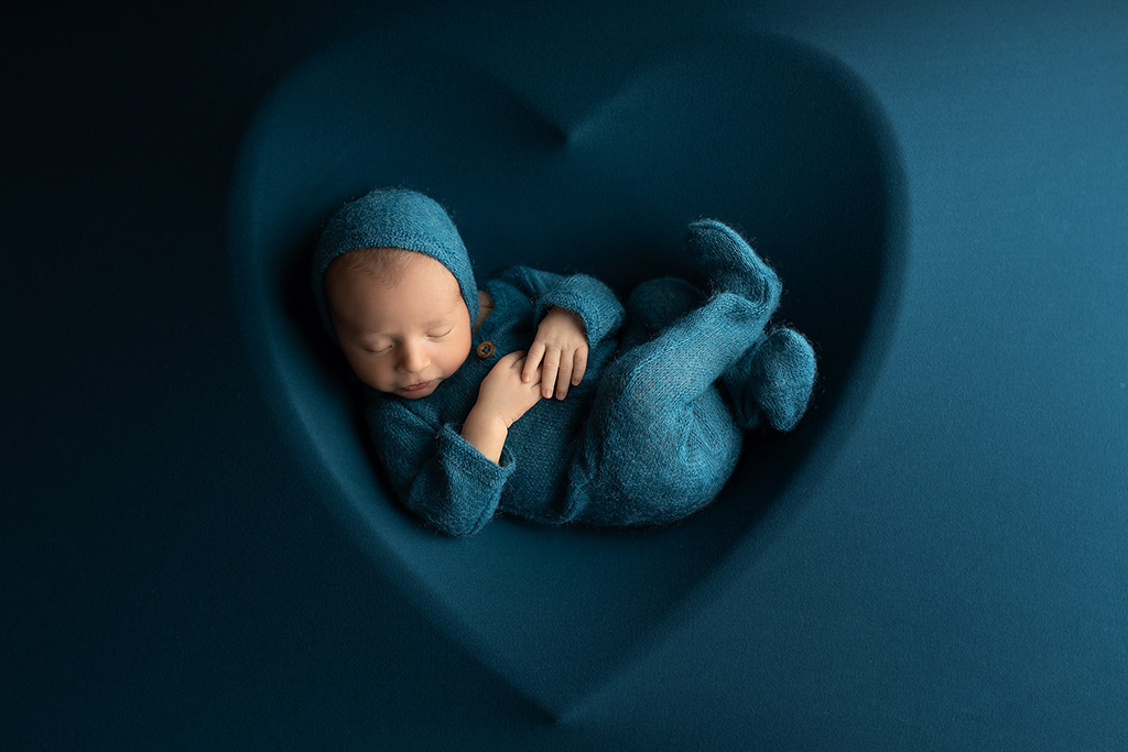 A peacefully sleeping newborn in a soft blue wrap, posed in a heart-shaped cradle backdrop.