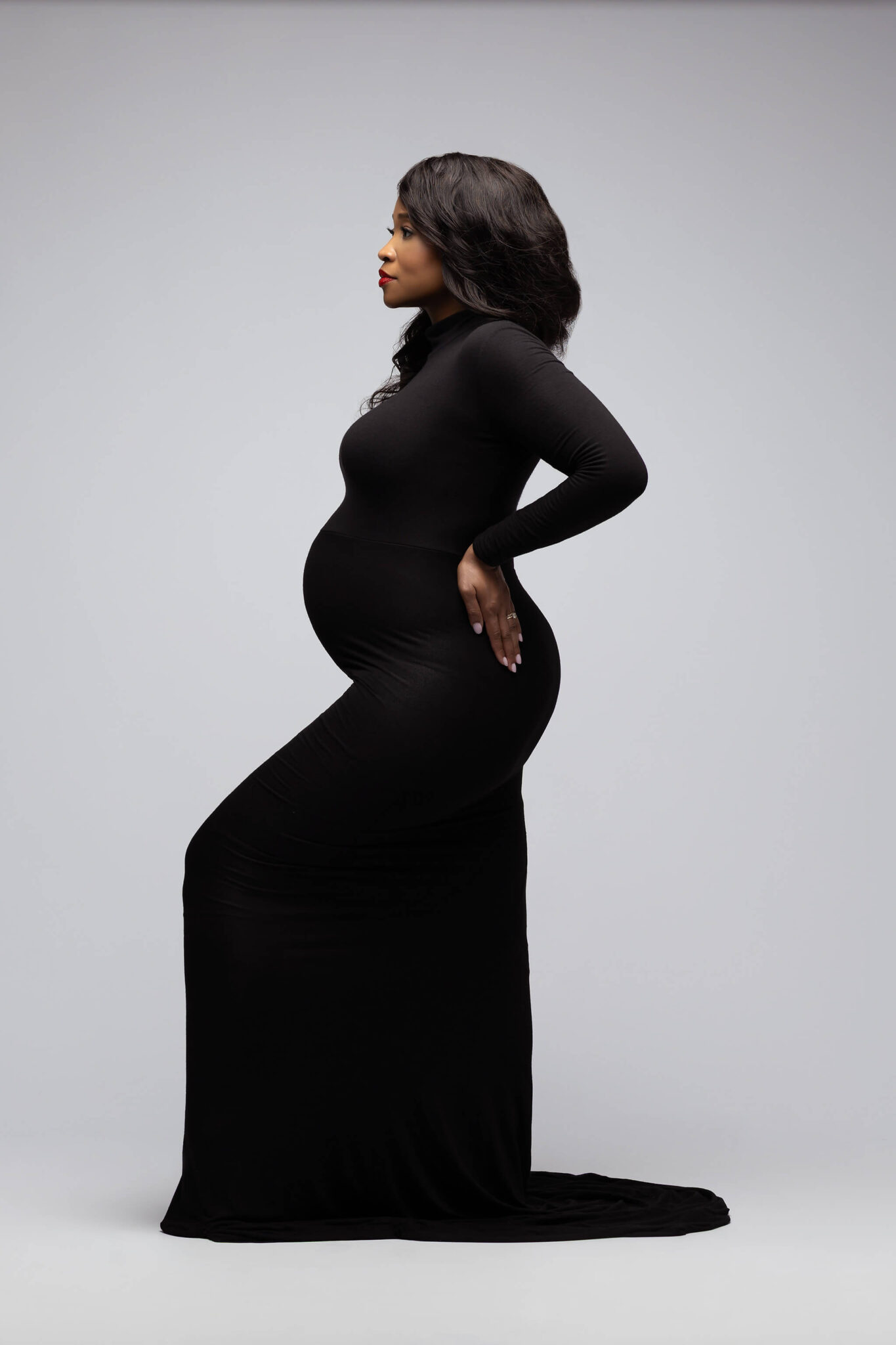 A mother to be in a black maternity gown stands with a hand on her hip in a studio in profile after meeting doulas in dallas