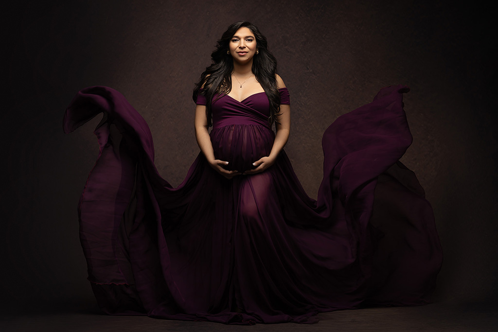 A pregnant woman in a flowing burgundy gown poses confidently in a studio portrait with dramatic fabric movement and rich tones, brought to you by Babymoon Concierge.