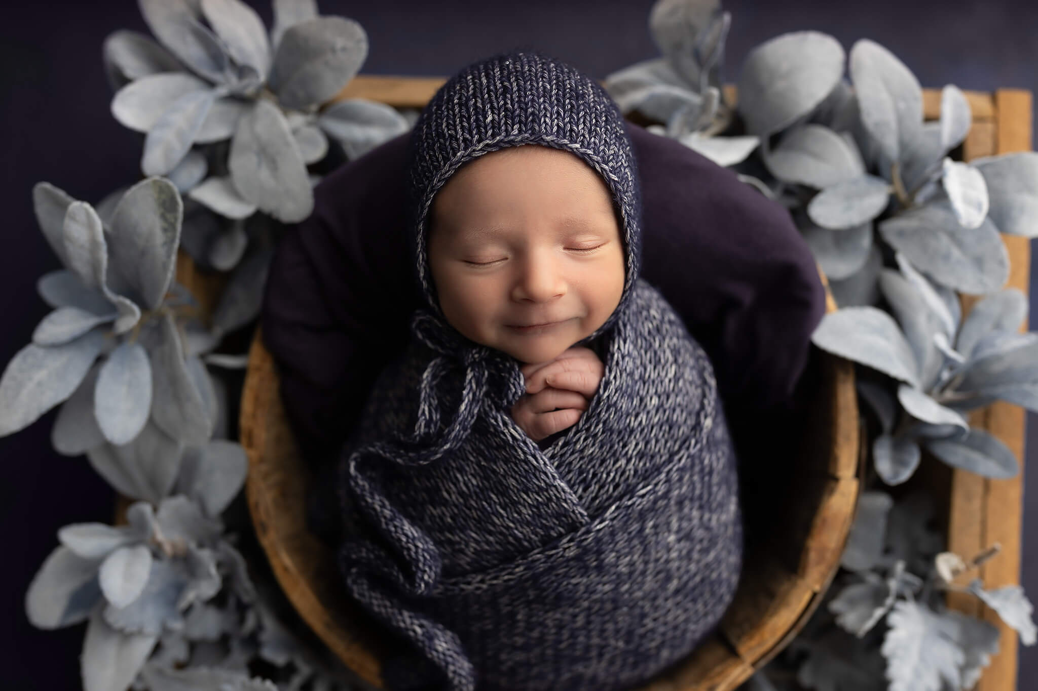 A smiling newborn wrapped tight in a knit swaddle and bonnet in a wooden bucket after visiting infant daycares in fort worth