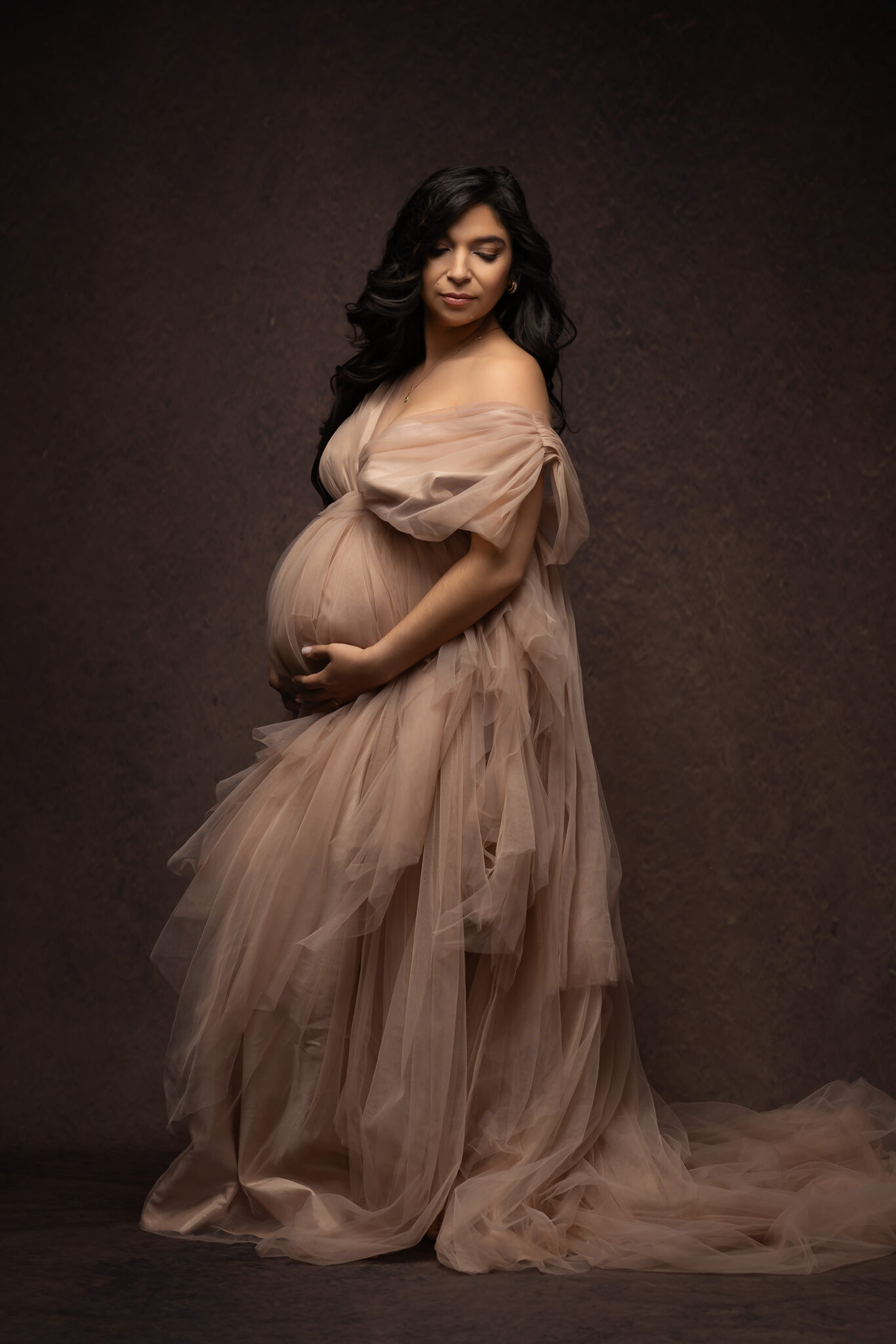 A pregnant woman gazes down her shoulder while standing in a pink tule maternity dress in a studio after meeting midwives in fort worth