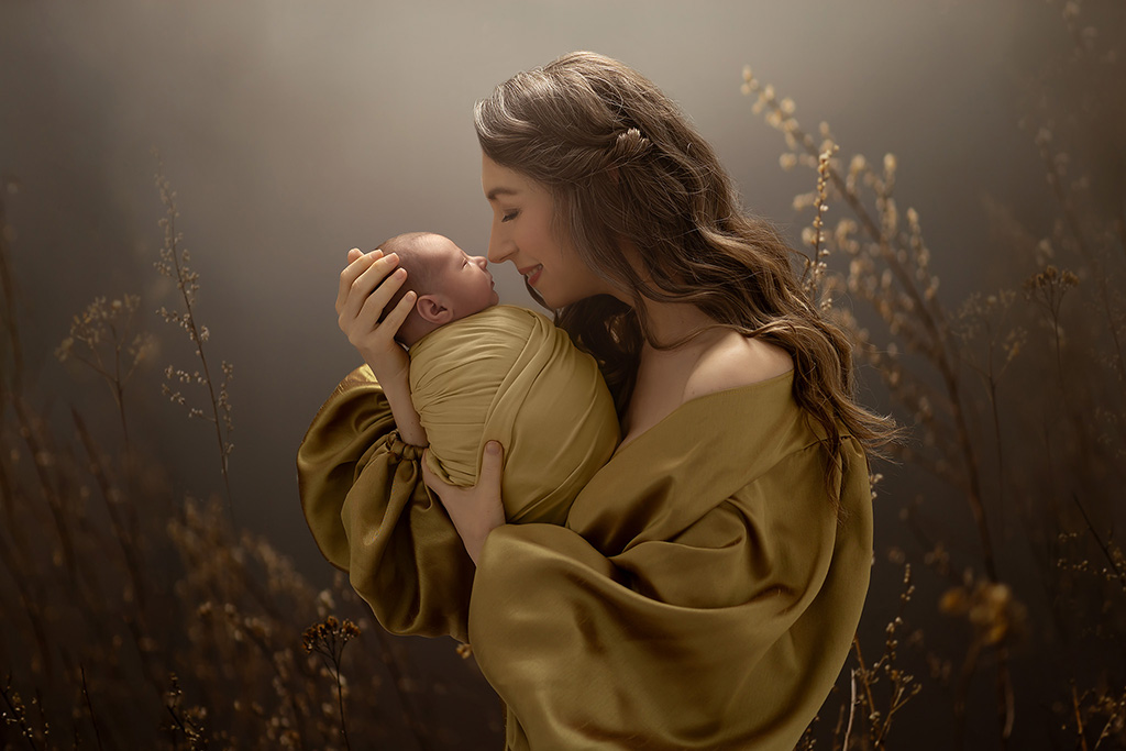 A mother in a golden dress lovingly holds her newborn wrapped in soft yellow, surrounded by warm light and tall grasses.