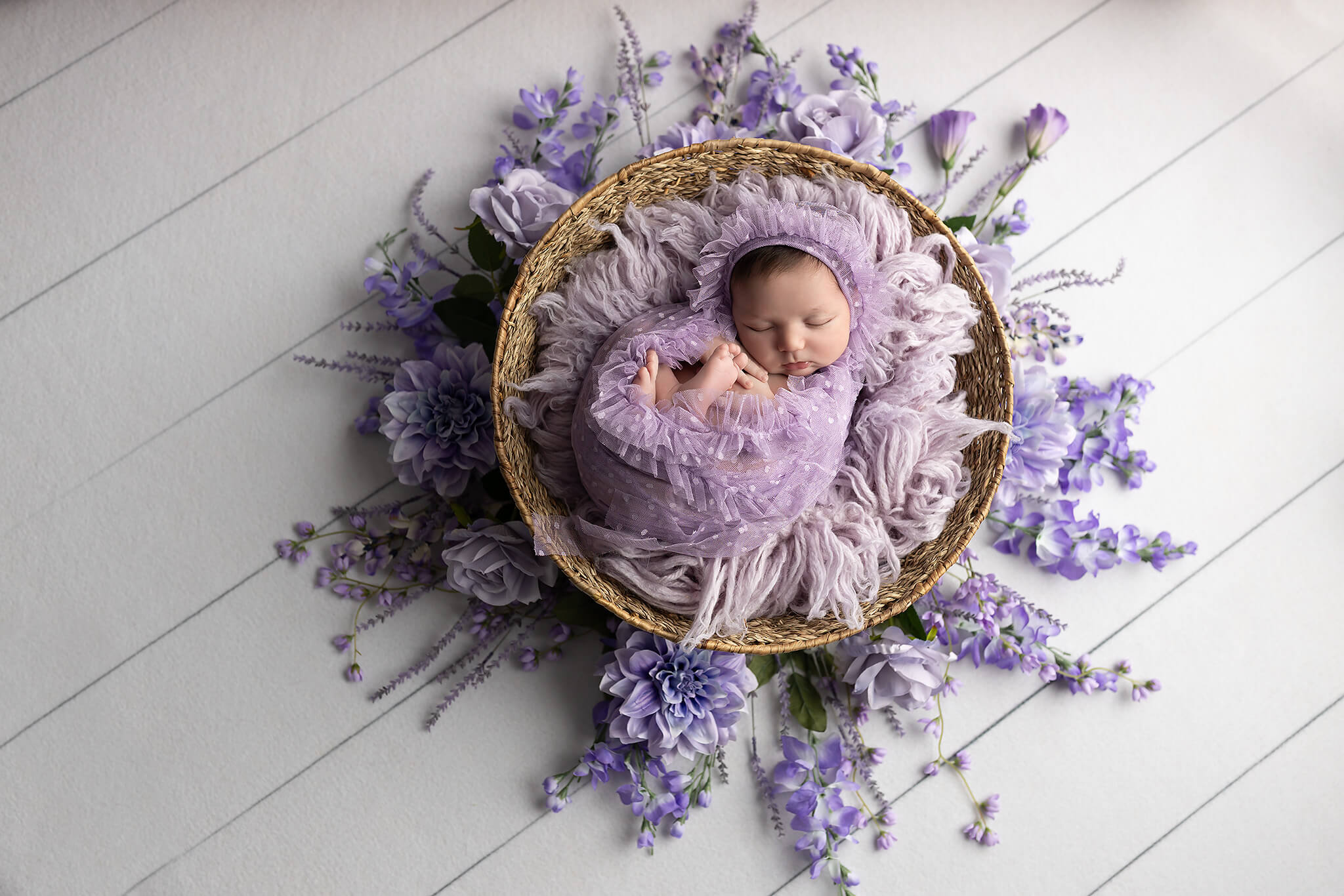 A sleeping newborn baby girl in a purple swaddle in a woven basket surrounded by flowers after meeting pediatric dentists in fort worth