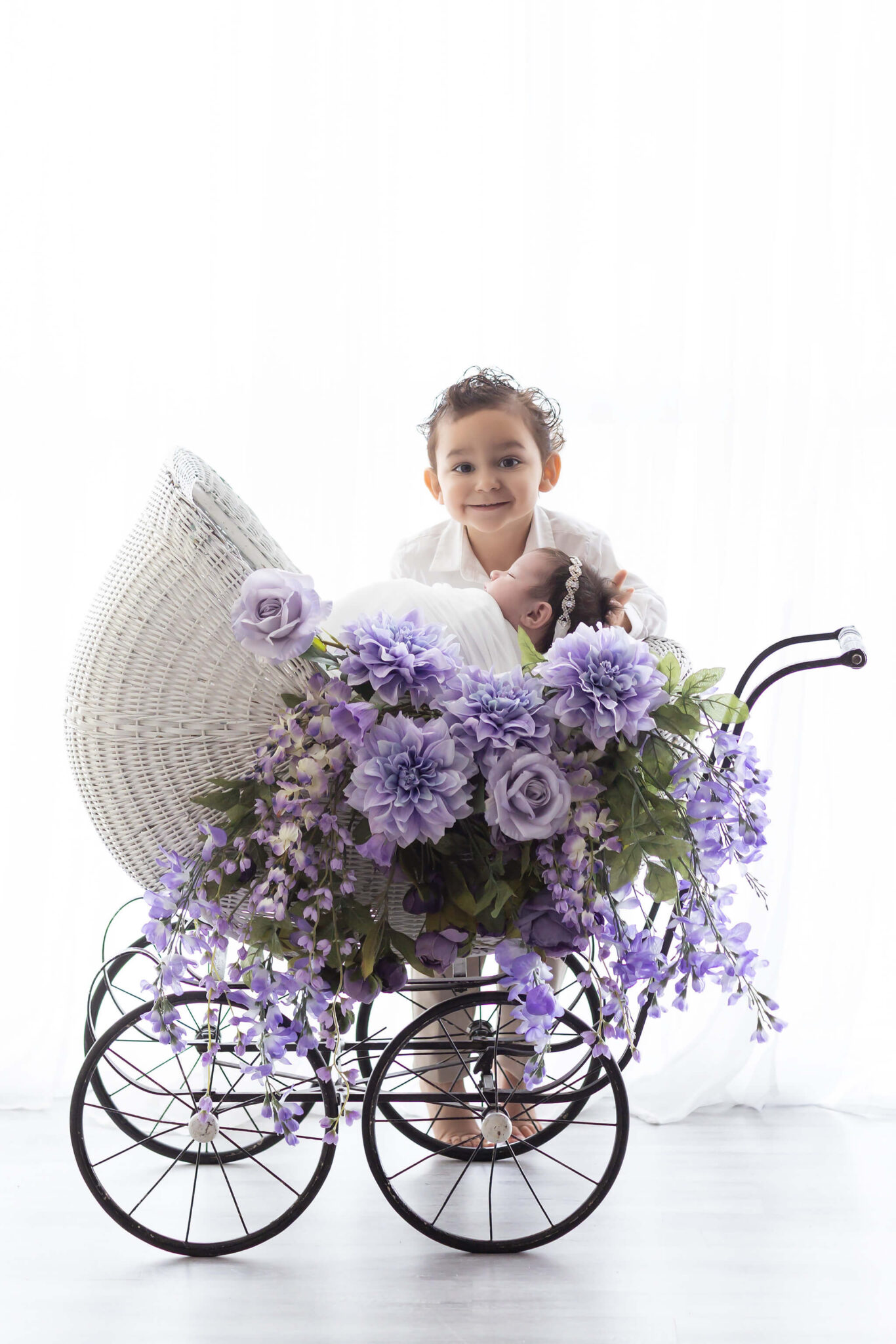 A toddler smiles big while standing behind his sleeping newborn sister in a flower covered baby carriage after meeting pediatric dentists in fort worth
