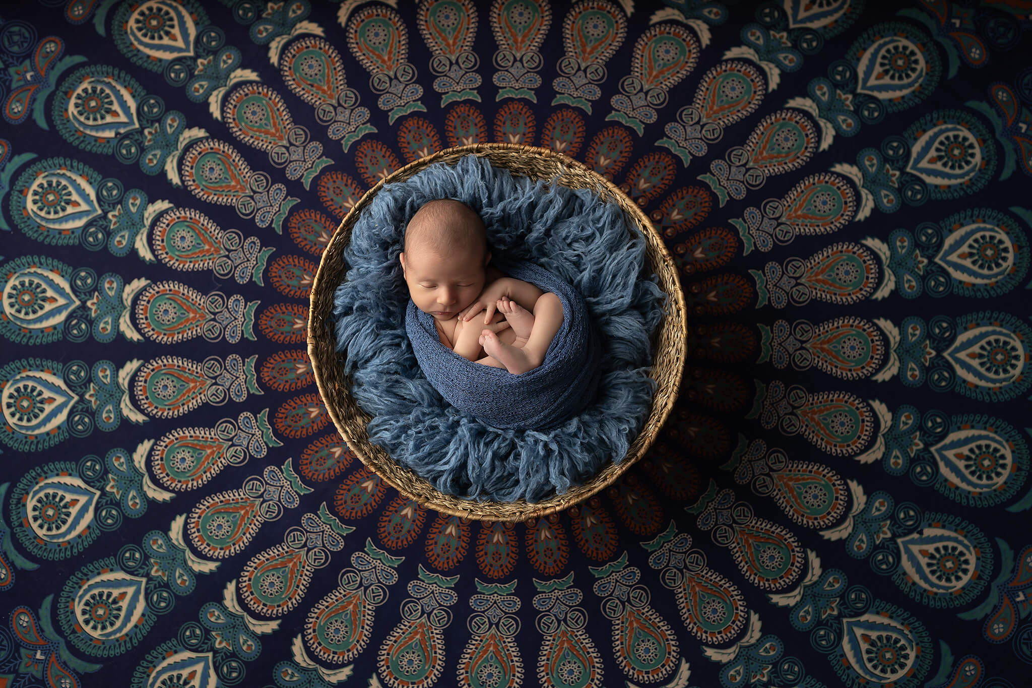 A newborn baby sleeps in a blue swaddle in a basket on an ornate round rug after meeting pediatricians in dallas