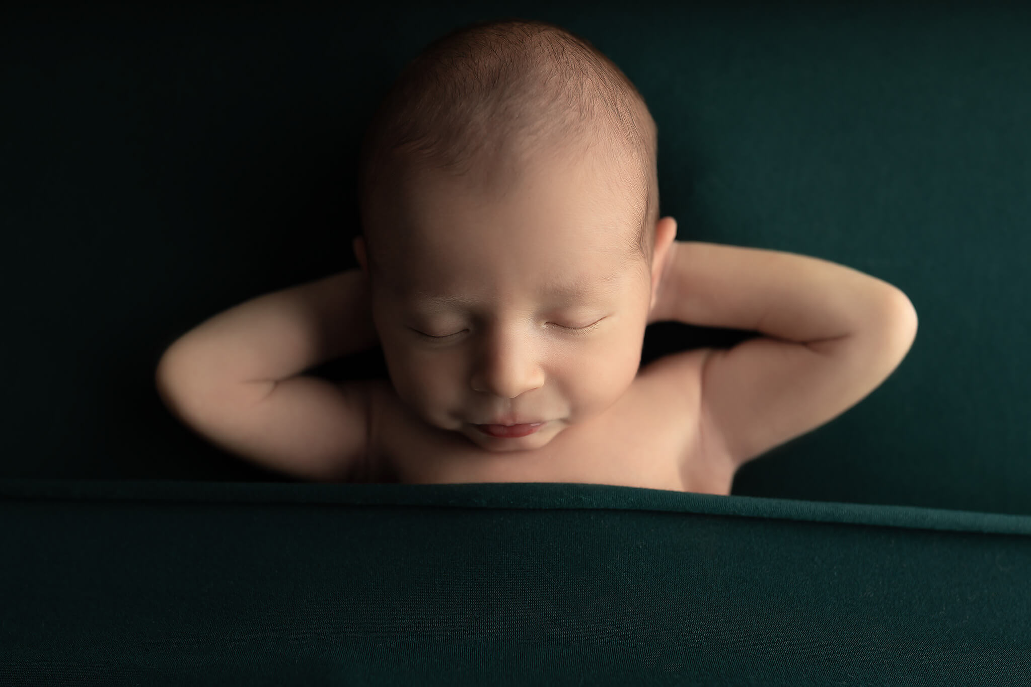 A newborn baby sleeps with hands behind its head on a forest green bed after meeting pediatricians in dallas