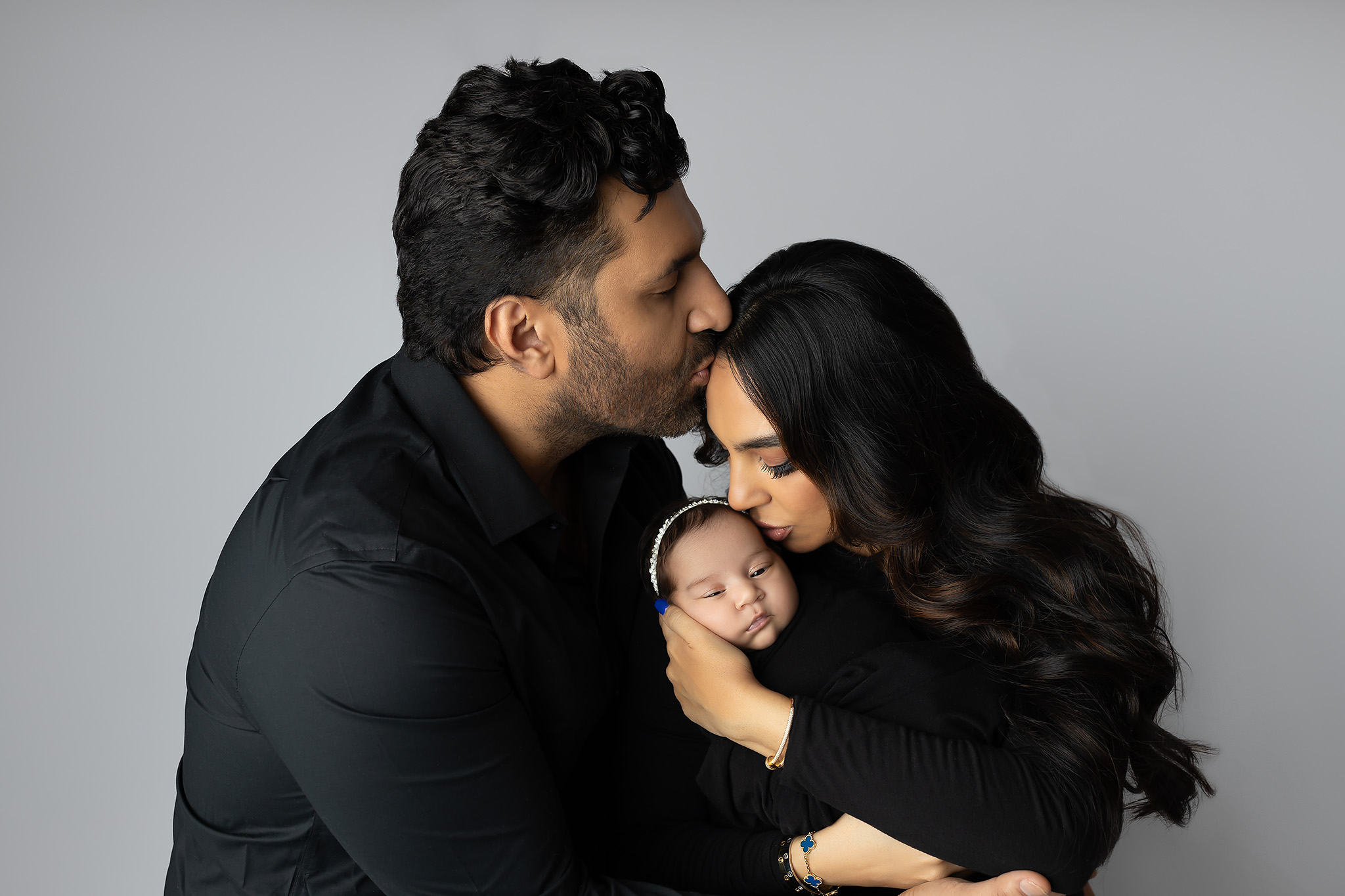 A newborn baby girl in a black swaddle is kissed on the head by mom as dad kisses her forehead after meeting postpartum doulas in dallas