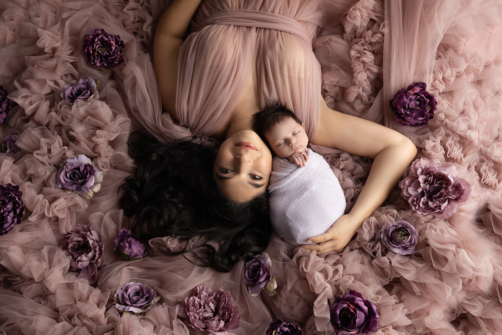 A mother to be smiles while laying on a bed of her tule maternity gown and flowers snuggling her sleeping newborn on her shoulder thanks to postpartum doulas in dallas