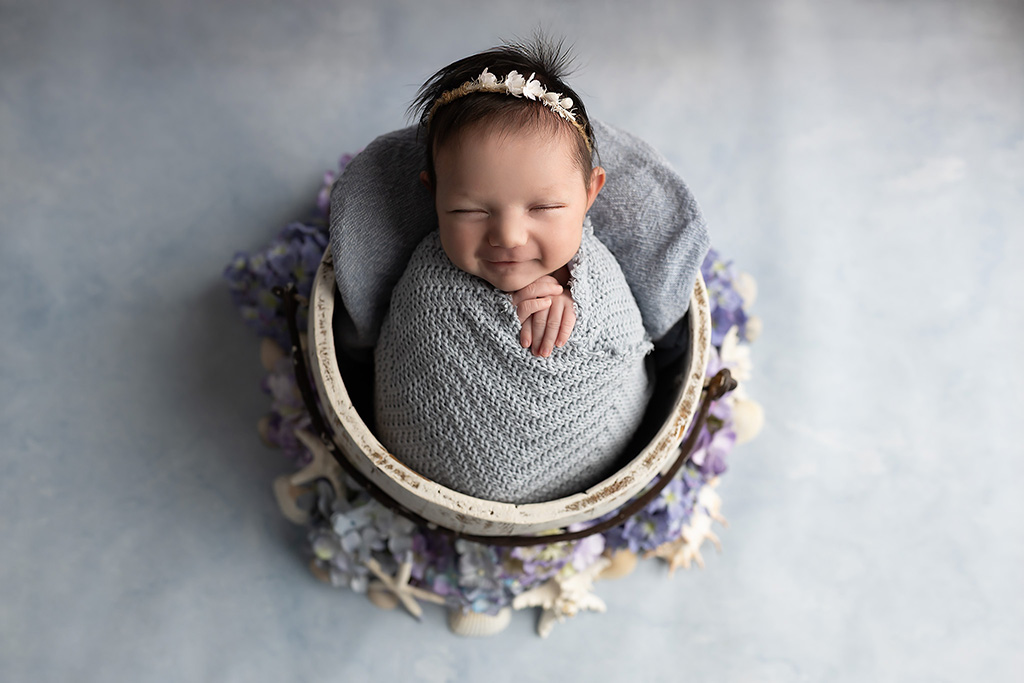 A smiling newborn wrapped in a soft gray knit blanket, posed in a round basket with purple and white floral accents on a soft blue background.