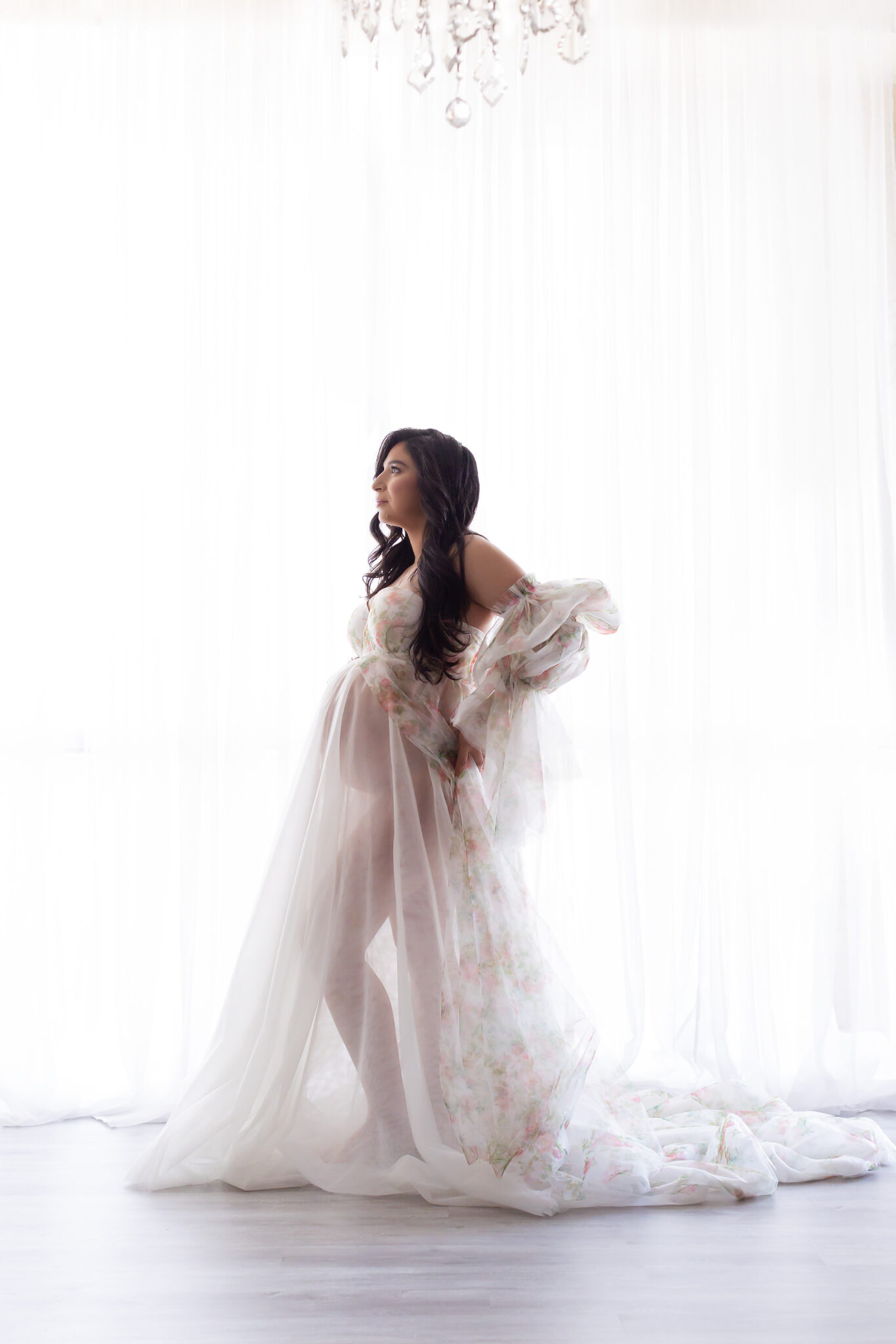 A pregnant woman in a sheer white maternity gown stands in a studio window with hands on hr back after finding baby shower venues in dallas