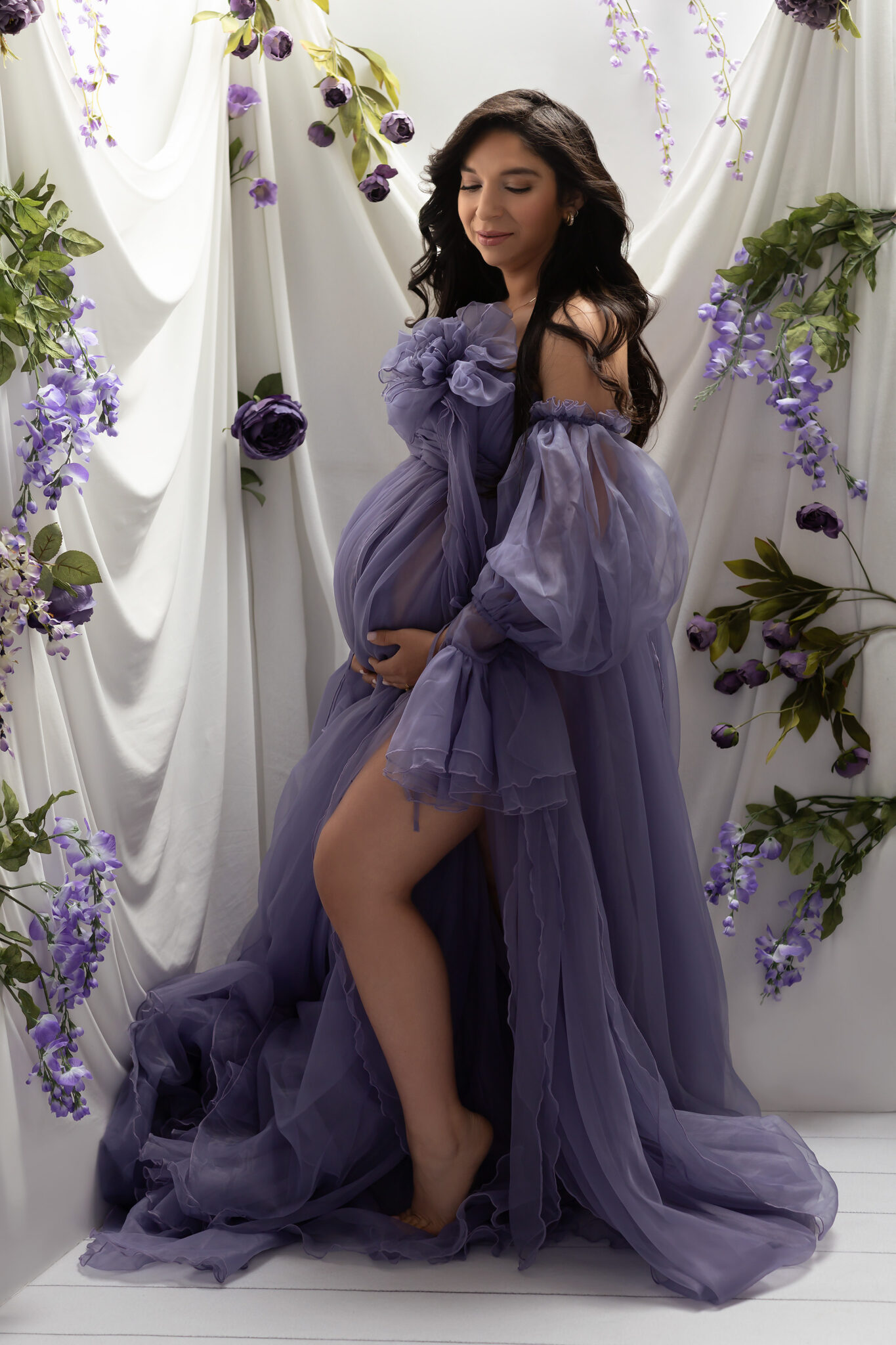 A mom to be stands in a studio in a purple tule dress gazing down her shoulder with hands under the bump after visiting baby shower venues in dallas