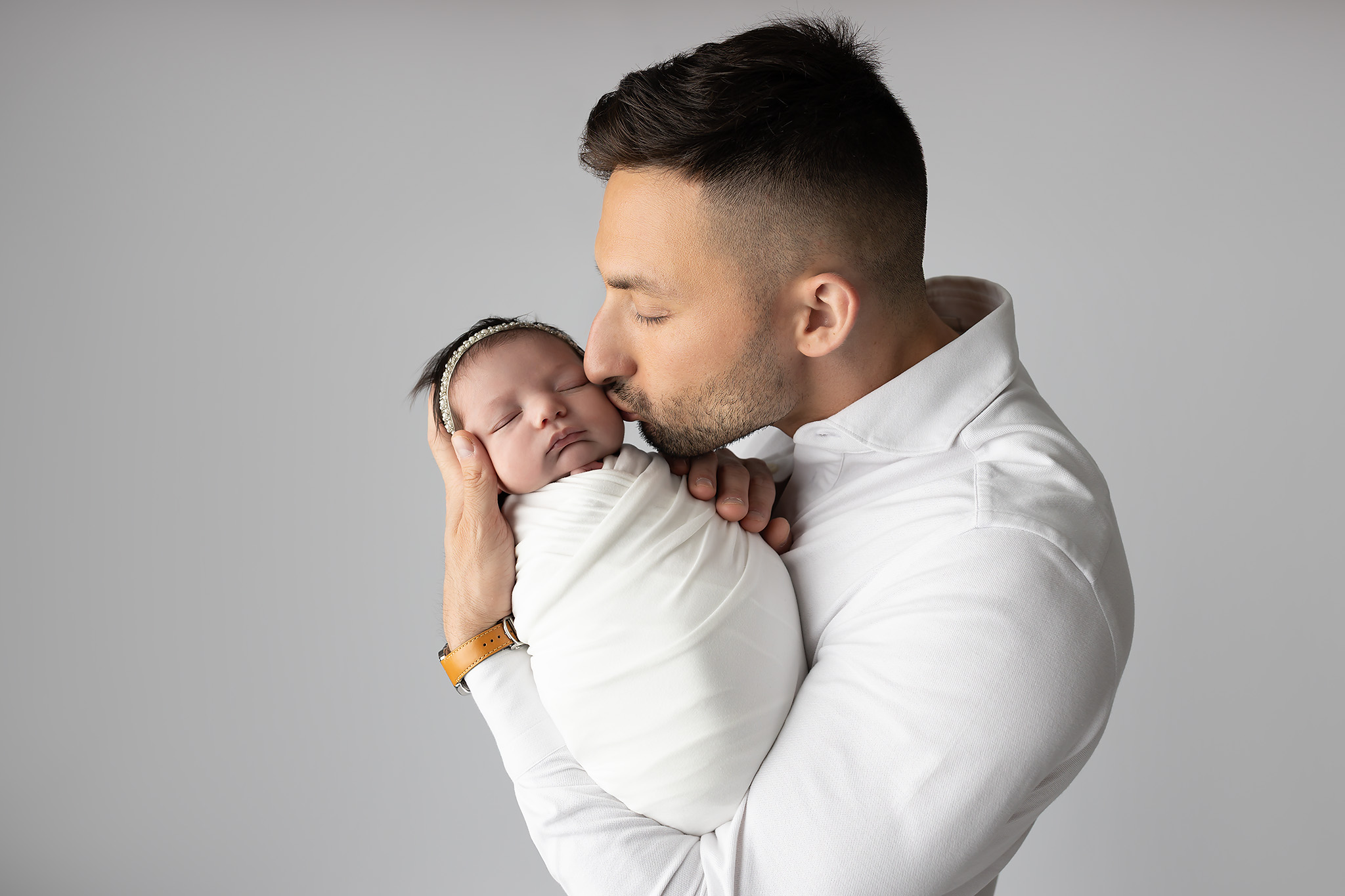 A happy dad kisses his sleeping newborn daughter in her arms while standing in a studio