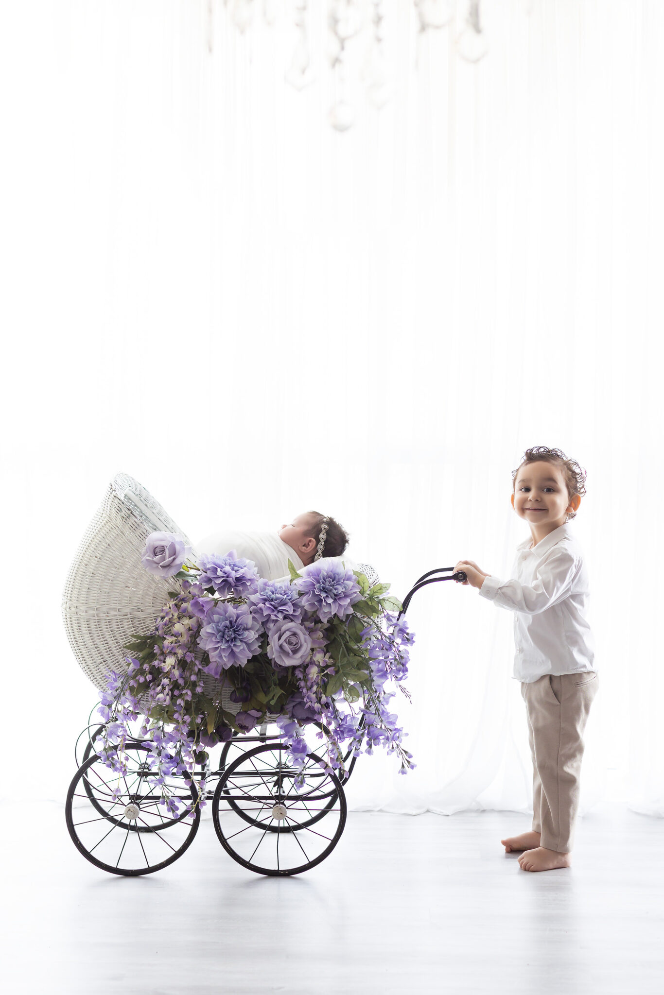 A toddler boy in a white shirt pushes a baby carriage holding his newborn sister and purple flowers in a studio after finding childcare in fort worth