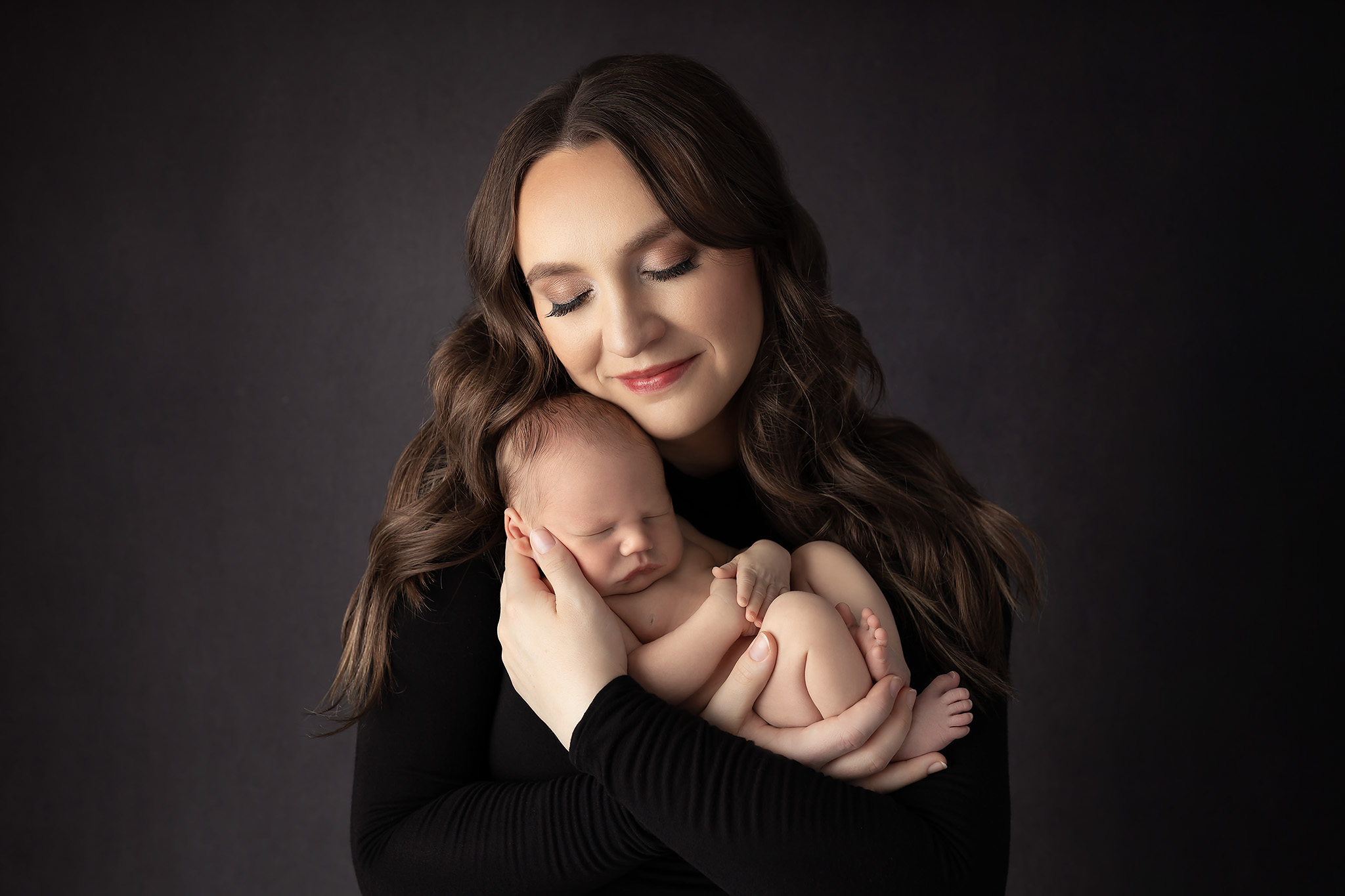 A new mom in a black long sleeve blouse snuggles her sleeping newborn against her cheek