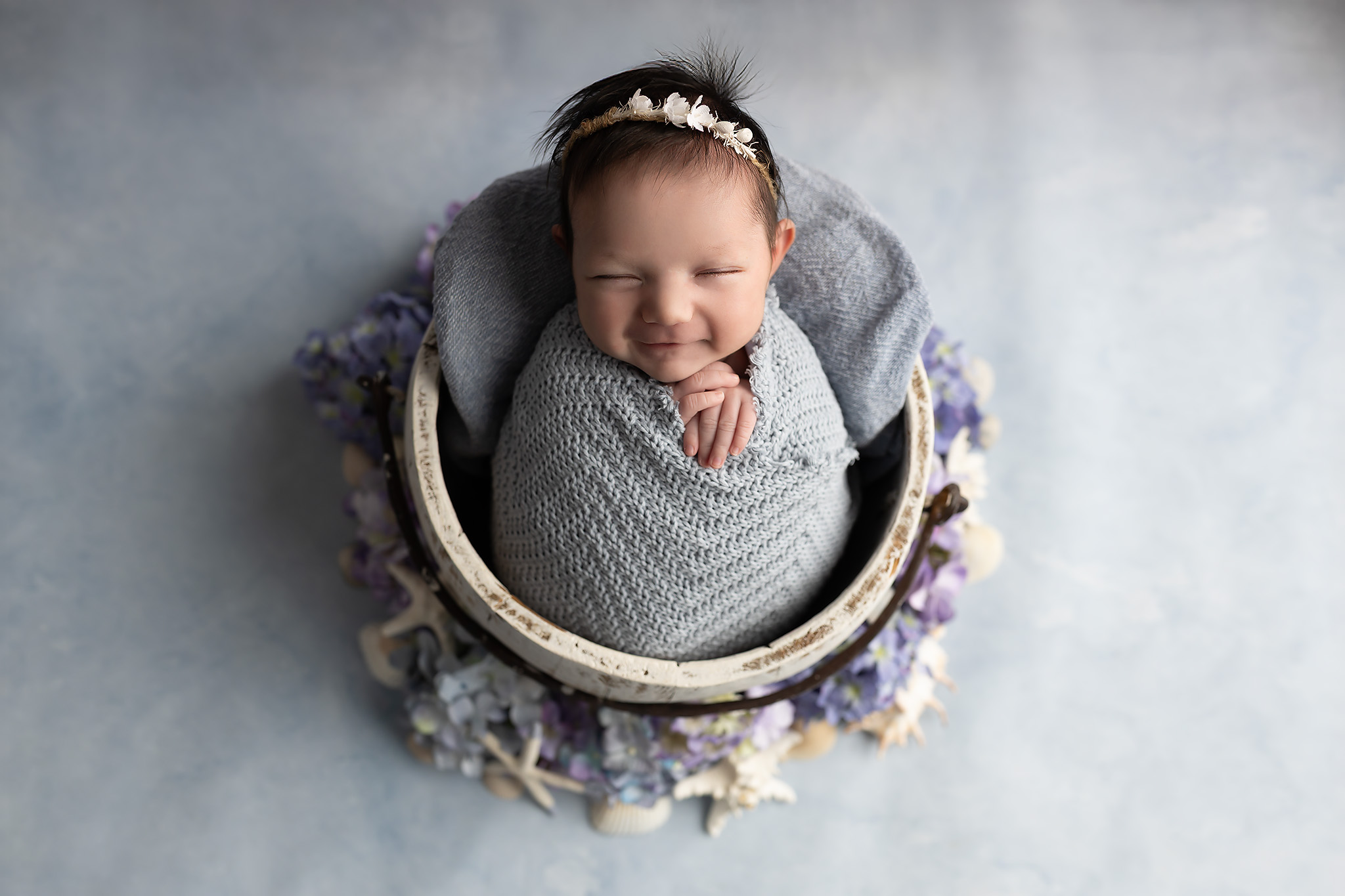 A newborn baby girl smiles in her sleep in a knit swaddle inside a wooden bucket after visiting pediatric dentists in dallas