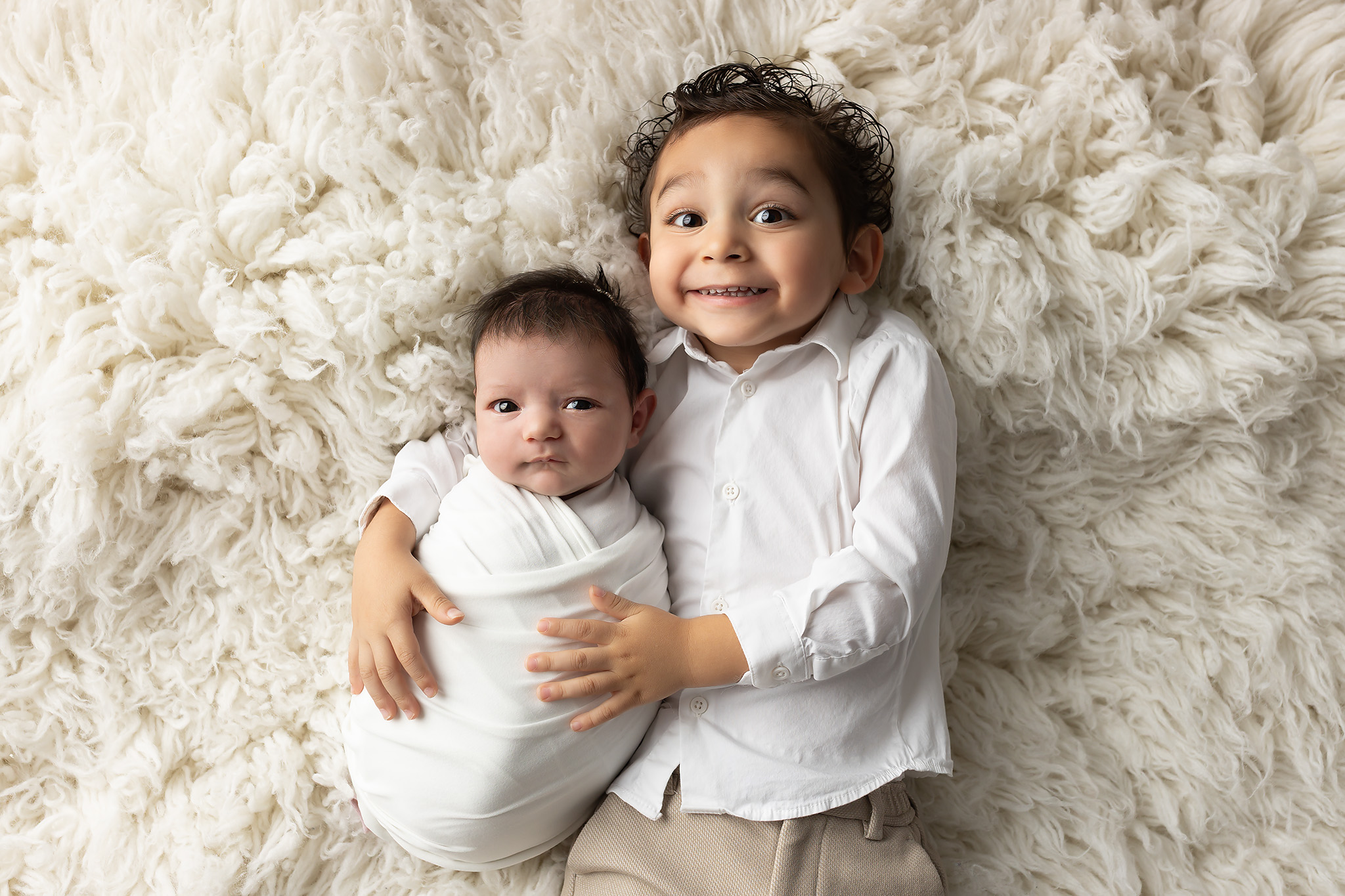 A toddler boy smiles big while laying on a fur blanket in a white shirt hugging his newborn baby sibling in a white swaddle with eyes open