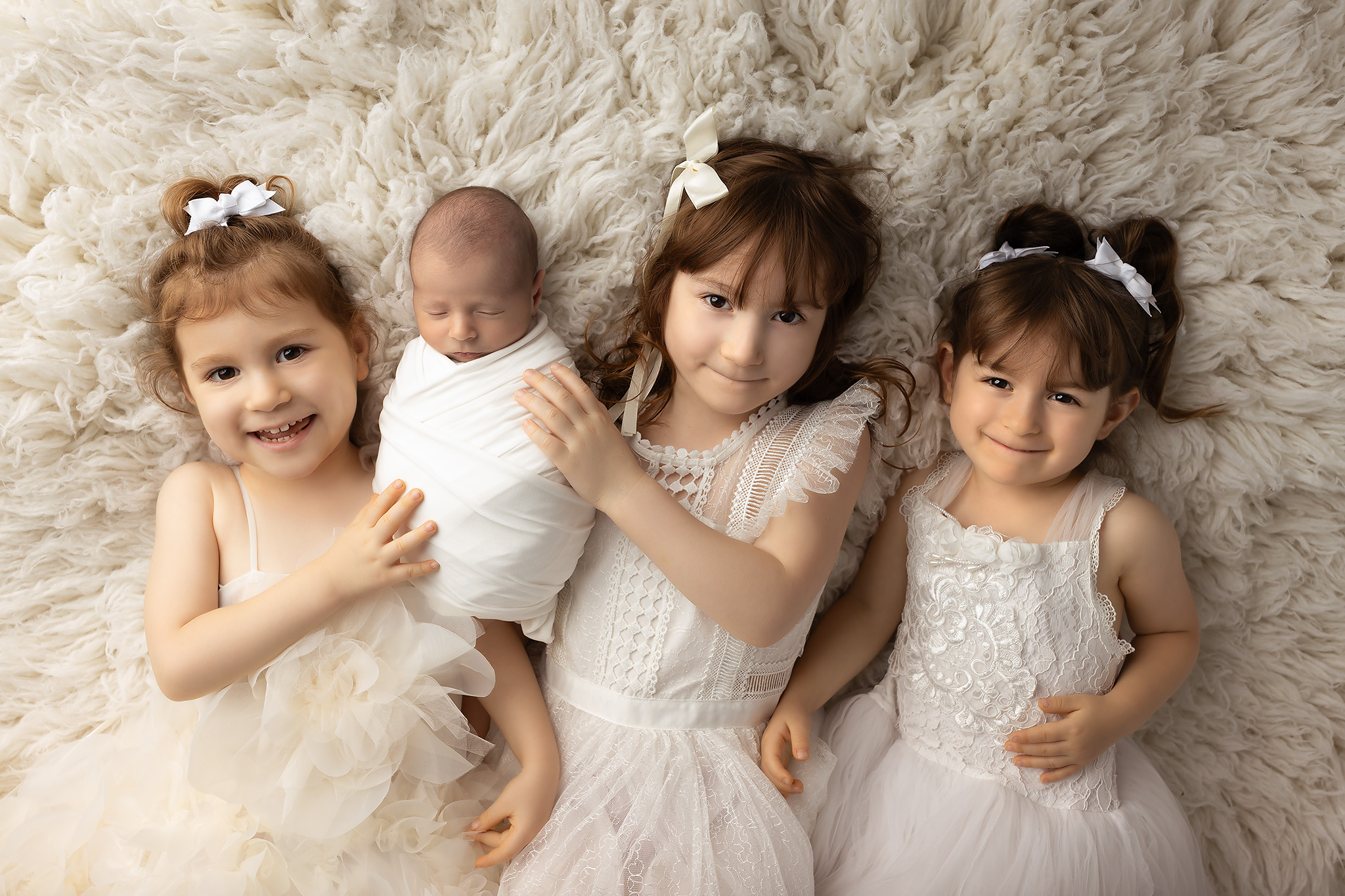 Three toddler girls in white dresses lay on a fur blanket with their sleeping newborn sibling in a white swaddle after visiting pediatricians in fort worth