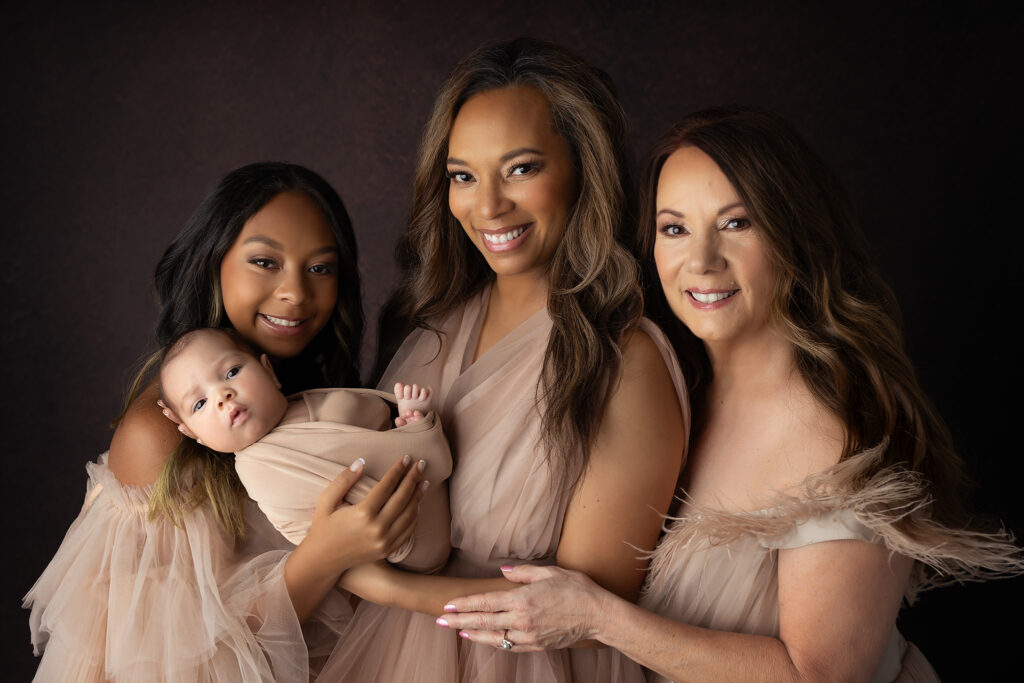 Three generations of women smile together while holding a newborn baby, captured in a timeless family portrait by Colleyville newborn photographer Lindsay Walden, with best lash extensions Colleyville.