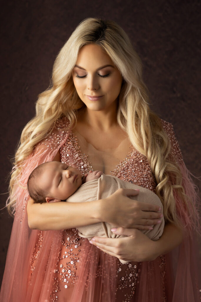 A mother in a blush pink gown cradles her newborn baby during a luxury newborn photography session in Colleyville.