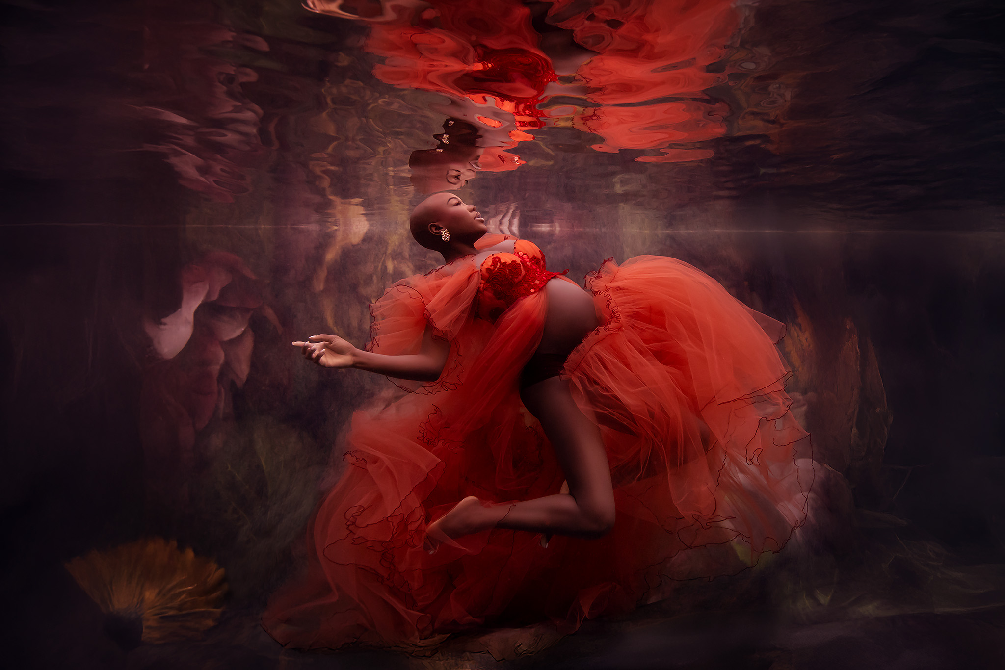 Beautiful dark skinned pregnant woman floating underwater in a an orange tulle gown with a floral backdrop