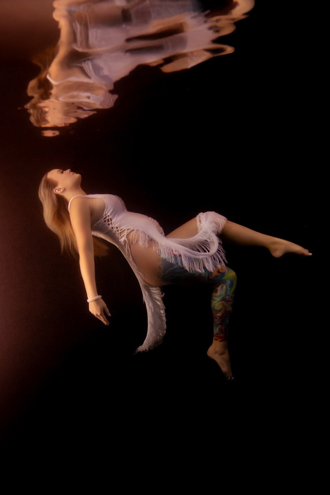 Underwater maternity image of woman in white fringe gown floating in front of a black backdrop