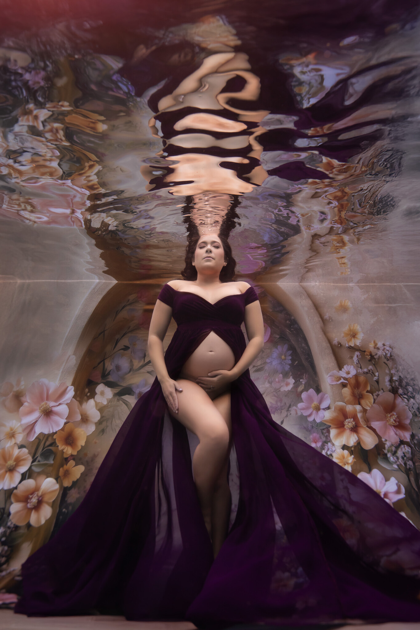 Serene underwater maternity session with pregnant woman in flowing purple gown open at the belly and surrounded by flowers