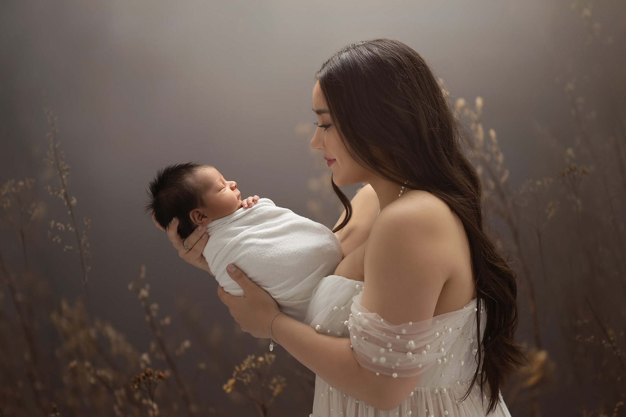 A luxury newborn photoshoot in Dallas with Lindsay Walden Photography showing a mother dressed in a white beaded gown holding her baby boy wrapped in white with beautiful light coming from behind.