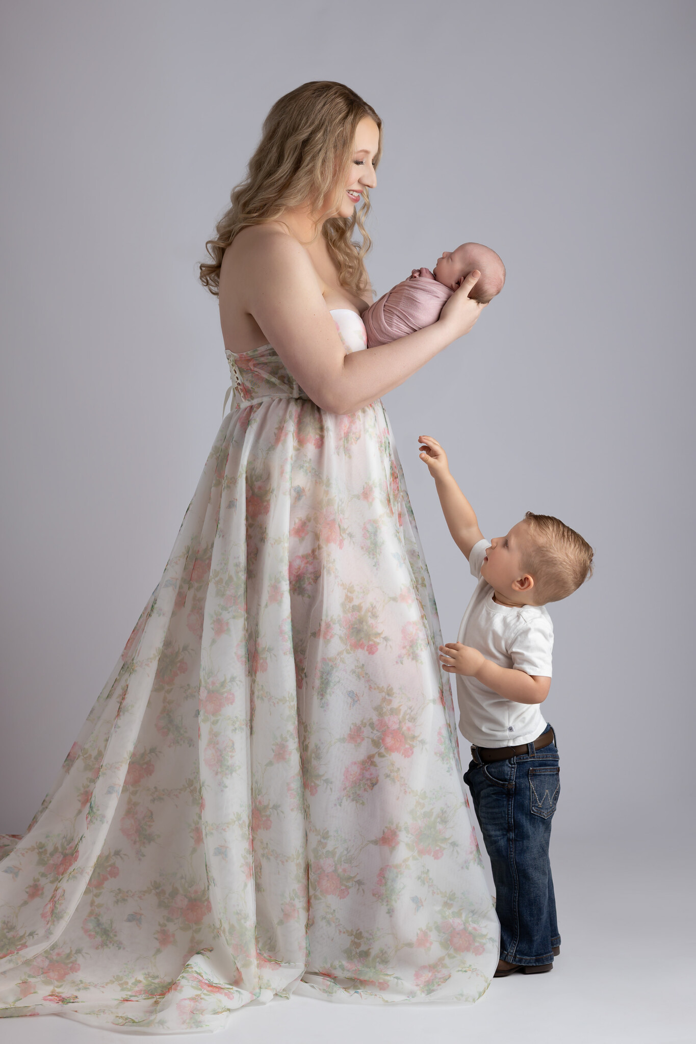 A mom holding her baby on her forearm while her toddler reaches up to gently touch his sibling. That is why moms should be in photos.