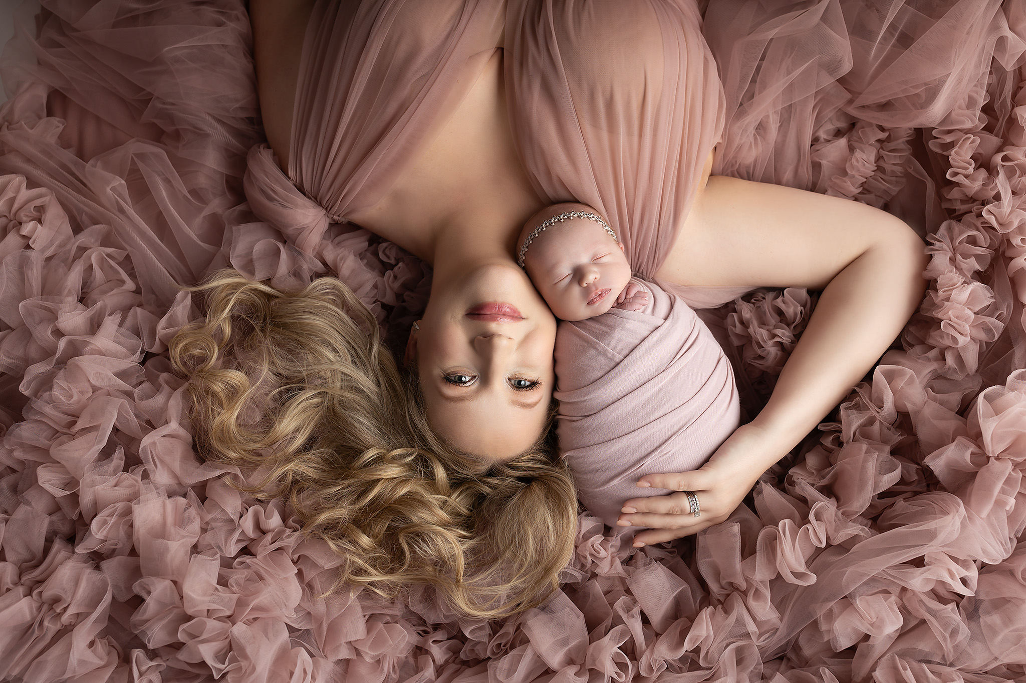A mother laying down on pink fluff with her baby resting in the nook between her shoulder and neck.
