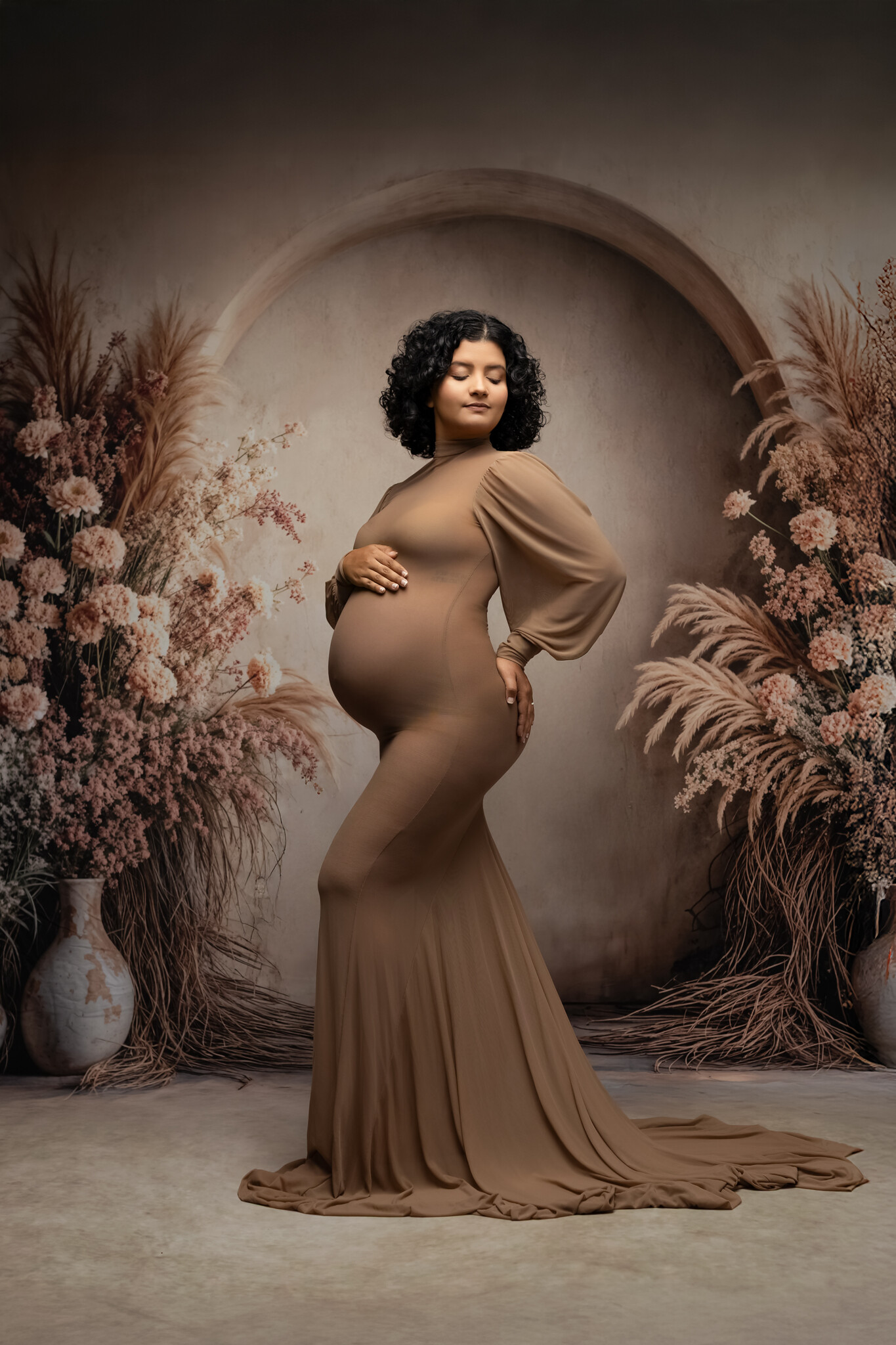 Gorgeous pregnant woman ready for her luxury baby shower in Dallas wearing a brown sheer gown surrounded by florals and an arched wall.