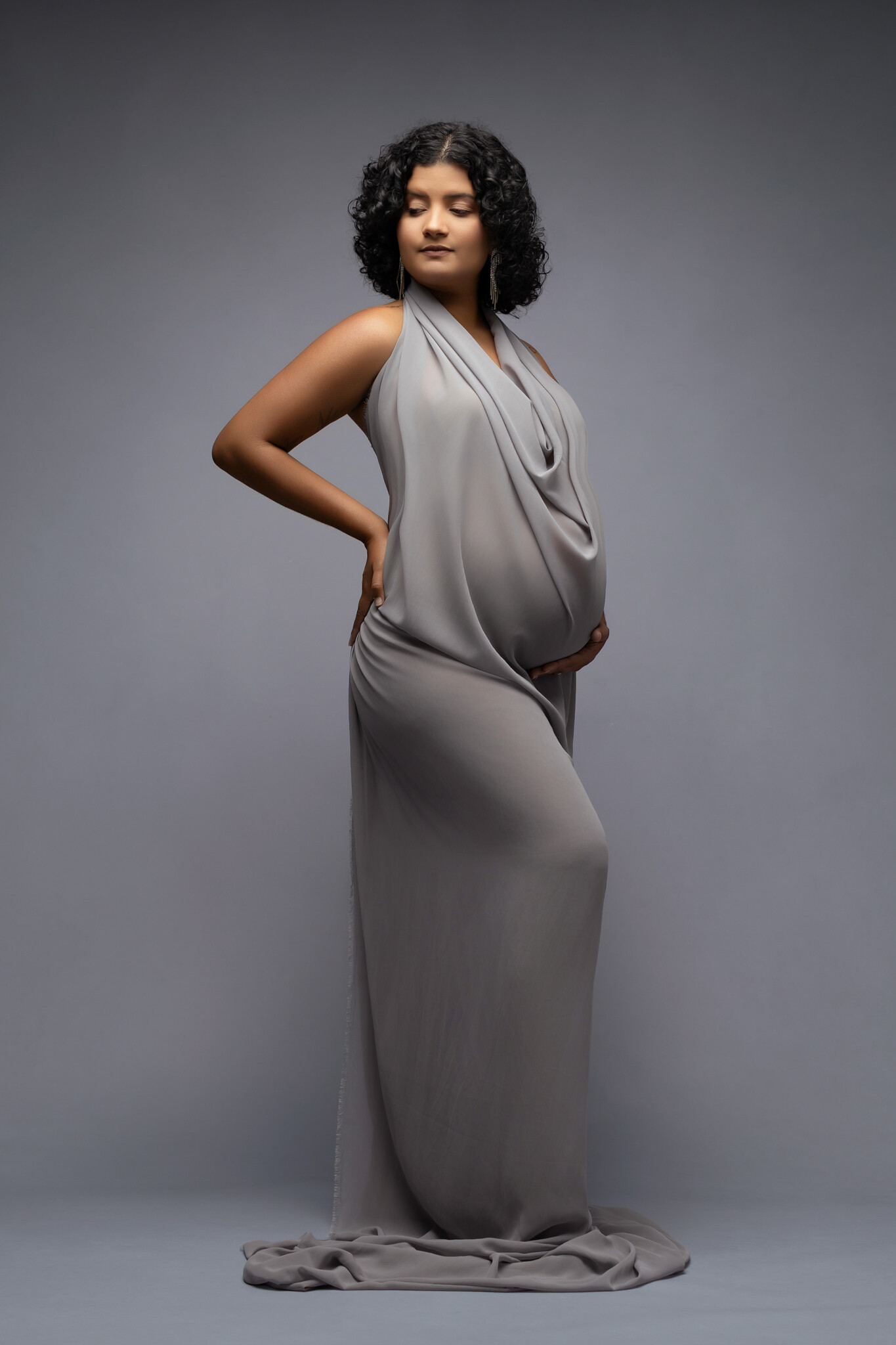 A monochromatic gray dress and backdrop with a gorgeous pregnant woman standing with a hand on her hip and one under her belly. The sereneness reflected in this image can be related back to Luxury Spas in Dallas for Expecting Moms.