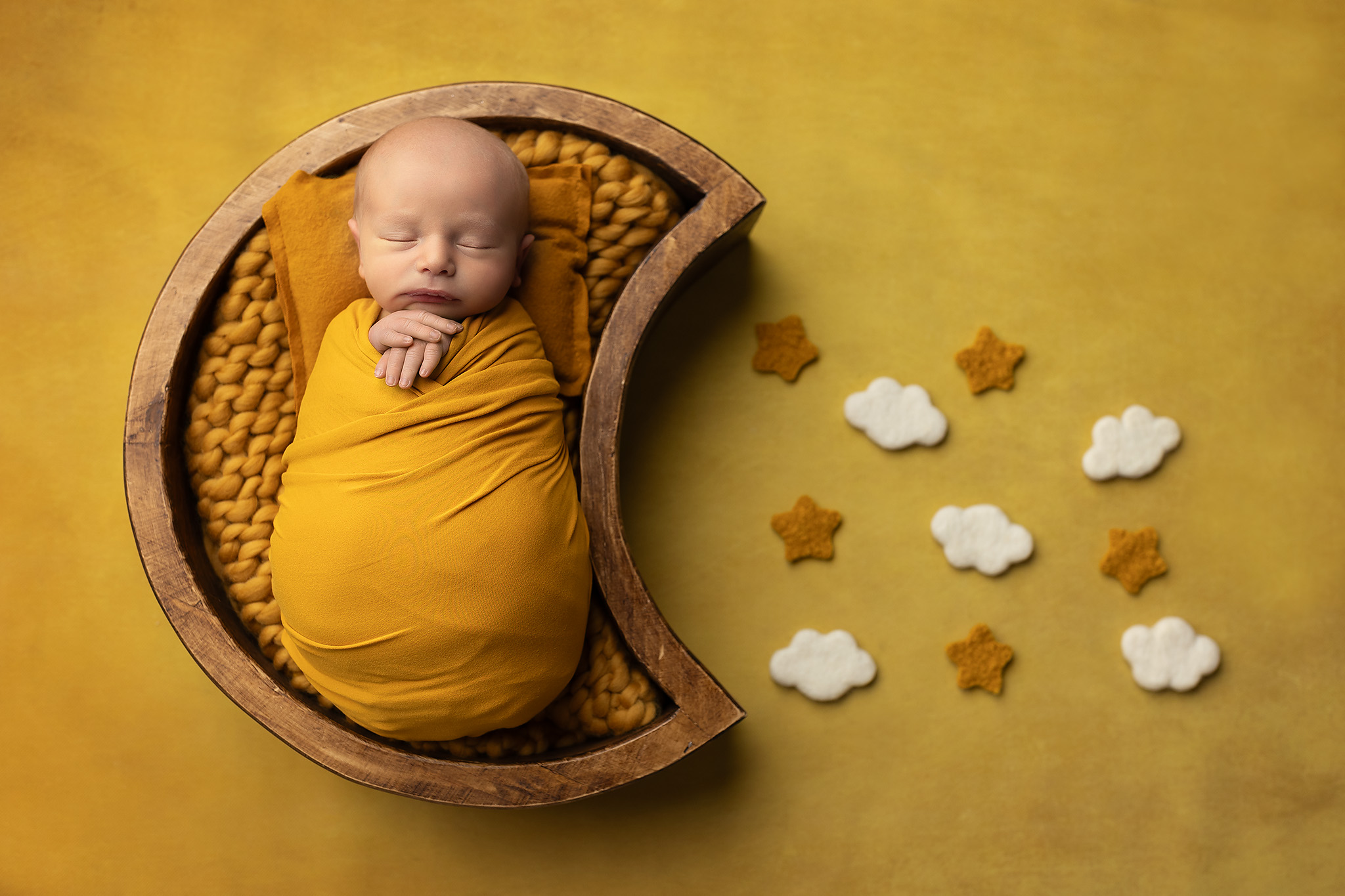 Newborn baby swaddled in yellow, laid in a moon prop with clouds and stars beside it. 
